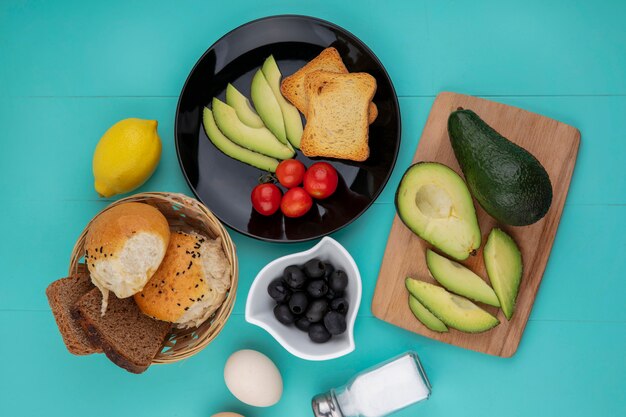 Top view of fresh avocados on wood kitchen board with a avocado slices tomatoes in a black plate with a bucket of breads on blue