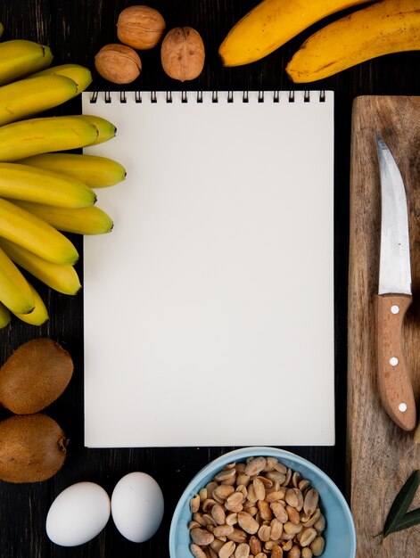 Top view of fresh banana and kiwi fruits with a sketchbook, walnuts and peanuts and kitchen knife on a wooden board on black