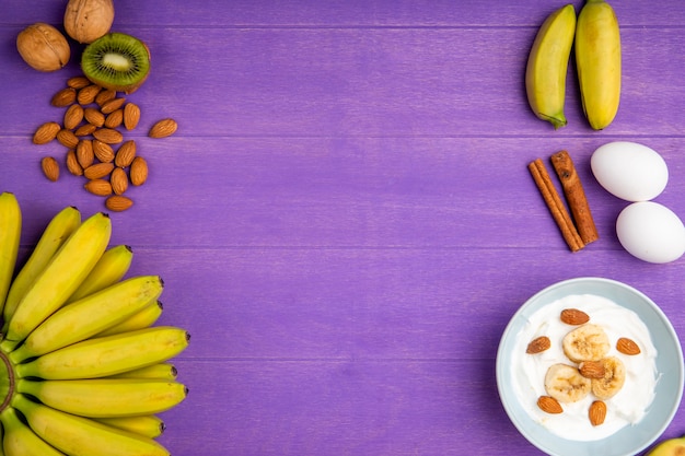 Top view of fresh bananas, cinnamon sticks, almond, two eggs and a bowl with yogurt and sliced bananas on purple wood with copy space