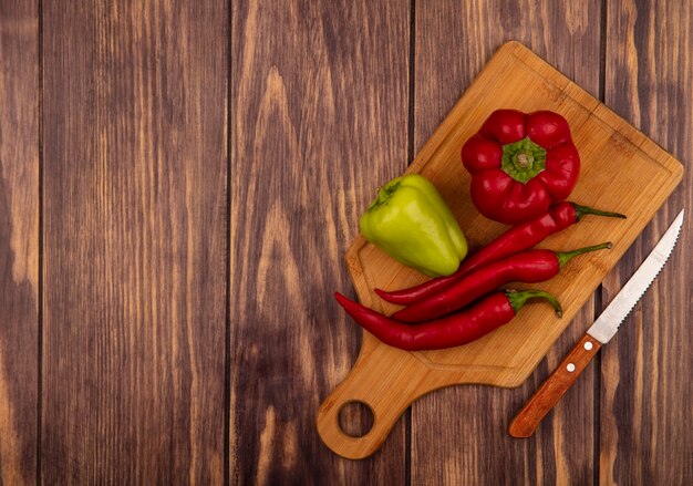 Top view of fresh bell and chili peppers on a wooden kitchen board with knife on a wooden wall with copy space