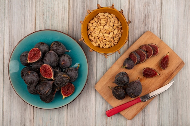 Top view of fresh black mission figs on a blue bowl with slices of black figs on a wooden kitchen board with knife with peanuts on a bucket on a grey wooden surface