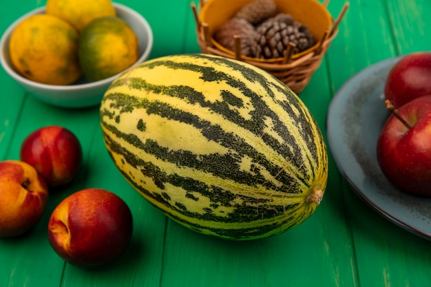 Top view of fresh cantaloupe melon with red apples on a plate with tangerines on a bowl with peaches isolated on a green wooden wall