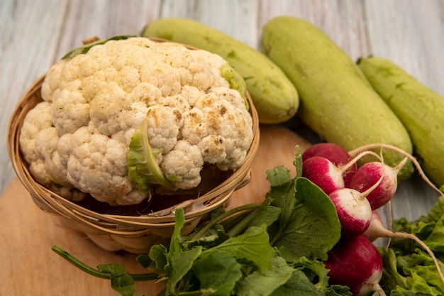 Vista dall'alto di cavolfiore fresco su un secchio su una tavola da cucina in legno con ravanelli e zucchine isolato su una superficie di legno grigia