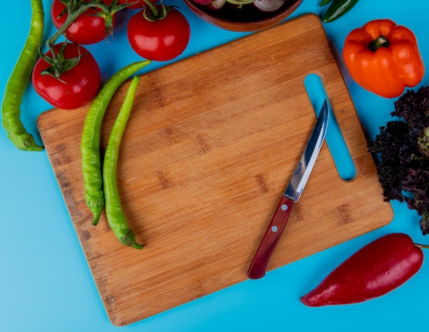 Top view of fresh chili peppers on a wood cutting board with kitchen knife and ripe tomatoes on blue