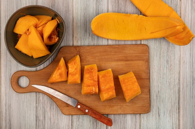 Top view of fresh chopped pumpkin slices on a wooden kitchen board with knife with pumpkin peels isolated on a grey wooden background