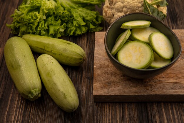 Top view of fresh chopped zucchinis on a bowl on a wooden kitchen board with lettuce zucchinis and cauliflower isolated on a wooden wall