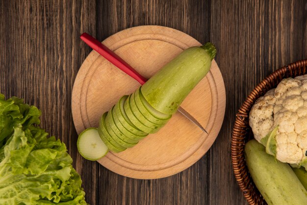 Top view of fresh chopped zucchinis on a wooden kitchen board with knife with vegetables such as zucchinis and cauliflower on a bucket on a wooden background