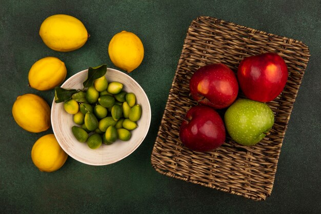 Top view of fresh colorful apples on a wicker tray with kinkans on a bowl with lemons isolated on a green surface