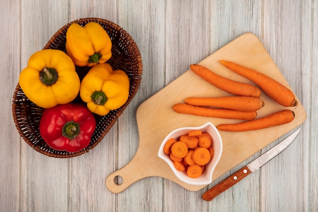 Top view of fresh colorful bell peppers on a bucket with carrots on a wooden kitchen board with knife on a grey wooden background