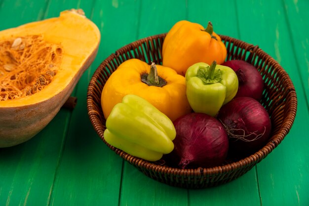 Top view of fresh colorful bell peppers on a bucket with red onion with pumpkin isolated on a green wooden wall