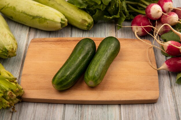 Top view of fresh cucumbers on a wooden kitchen board with radishes zucchinis and celery isolated on a grey wooden surface