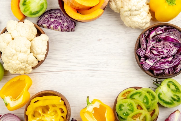 Top view fresh cut vegetables green tomatoes red cabbage onion pumpkin cauliflower yellow bell pepper in bowls on white wooden surface free space in center