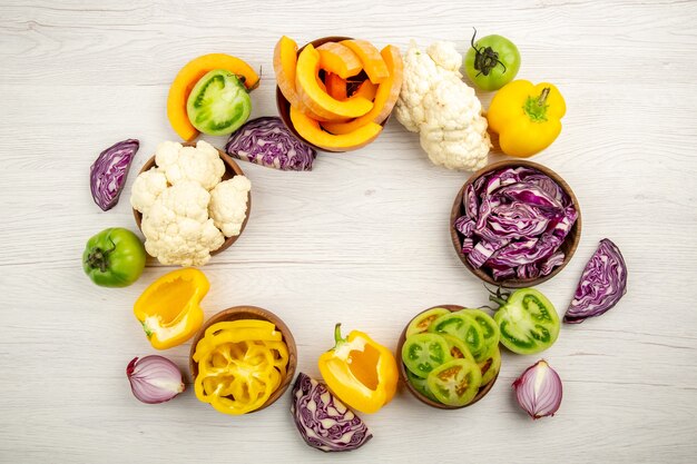 Top view fresh cut vegetables green tomatoes red cabbage onion pumpkin cauliflower yellow bell pepper in bowls on white wooden surface with free space