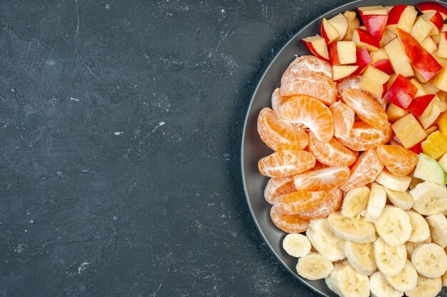 Вид сверху салат из свежих фруктов, нарезанные бананы, яблоки и апельсины на темном фоне
