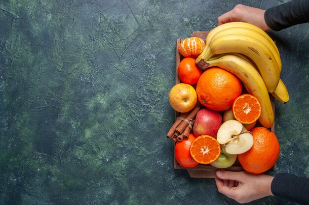 Top view fresh fruits oranges mandarins apples bananas and cinnamon sticks on wooden tray in female hands on dark background free space