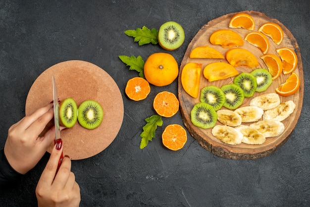 Free photo top view of fresh fruits set on a wooden tray and hand chopping kiwifruit on cutting board on black surface