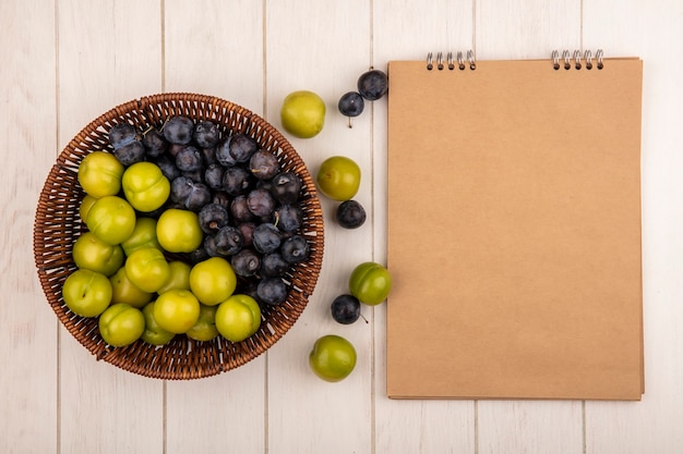 Top view of fresh fruits such as dark purple sloes and green cherry plums on a bucket on a white background with copy space