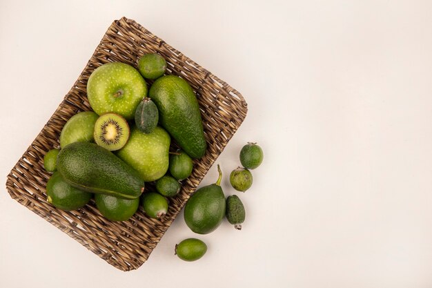 Top view of fresh fruits such as green apple feijoas avocados on a wicker tray on a white wall with copy space