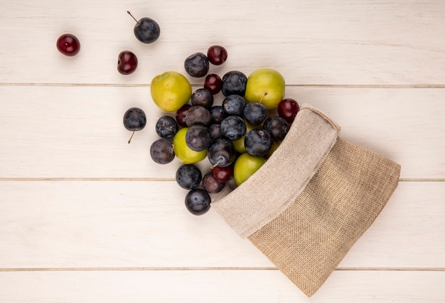 Top view of fresh fruits such as green cherry plumred cherries and sloes falling out of a burlap bag on a white wooden background