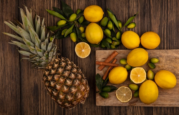 Top view of fresh fruits such as kinkans and lemons with cinnamon sticks on a wooden kitchen board with pineapples on a wooden surface
