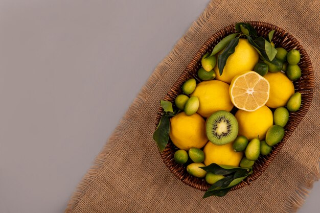 Top view of fresh fruits such as kiwi kinkans and lemons on a bucket on a sack cloth on a grey wall with copy space