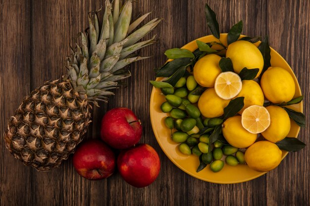 Top view of fresh fruits such as lemons and kinkans on a yellow dish with pineapple and red apples isolated on a wooden surface