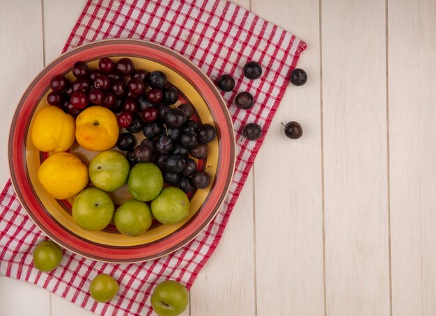 Top view of fresh fruits such as red cherriesgreen cherry plums and peaches on a bowl on a checked cloth on a white wooden background with copy space