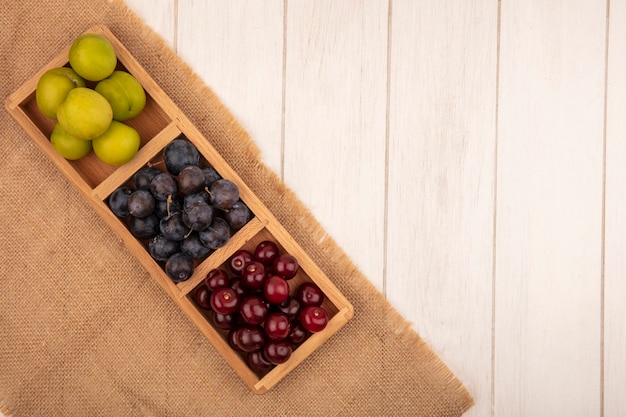 Top view of fresh fruits such as sloescherries and green cherry plum on a wooden divided tray on a sack cloth on a white wooden background with copy space