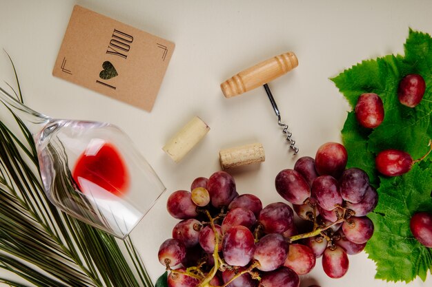 Top view of fresh grape , small postcard, bottle screw with wine corks and a wine glass lying on white table