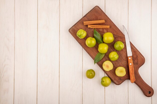 Top view of fresh green cherry plum on a wooden kitchen board with cinnamon sticks with knife on a white wooden background with copy space