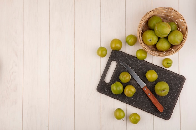 Top view of fresh green cherry plums on a kitchen cutting board with knife with cherry plums on a bucket on a white wooden background with copy space