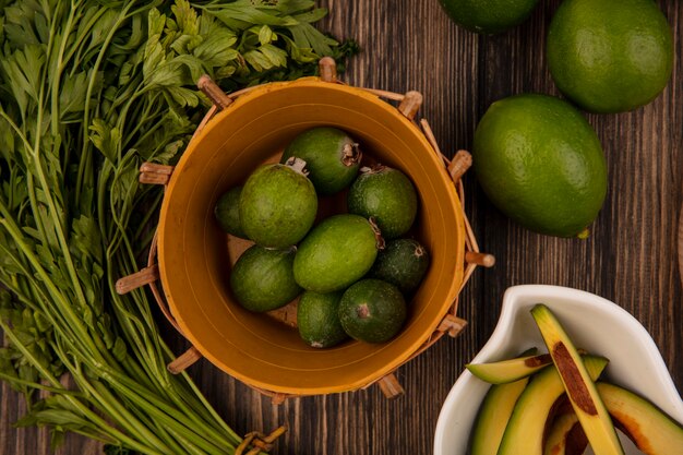 Top view of fresh green feijoas on a bucket with avocado slices on a bowl with limes and parsley isolated on a wooden background