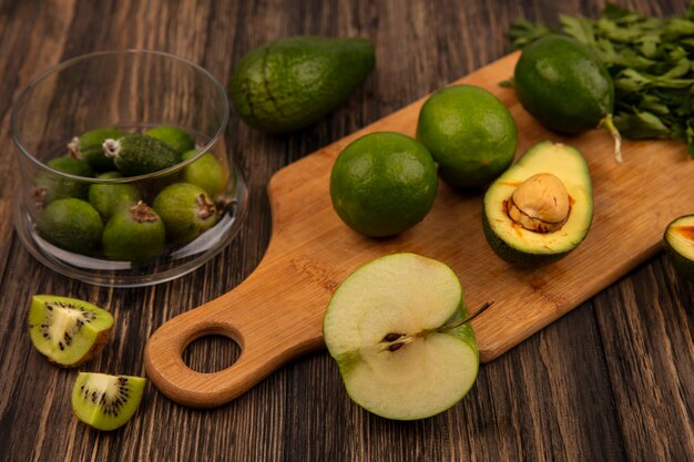 Top view of fresh green limes on a wooden kitchen board with feijoas on a glass bowl with avocados kiwi and parsley isolated on a wooden wall