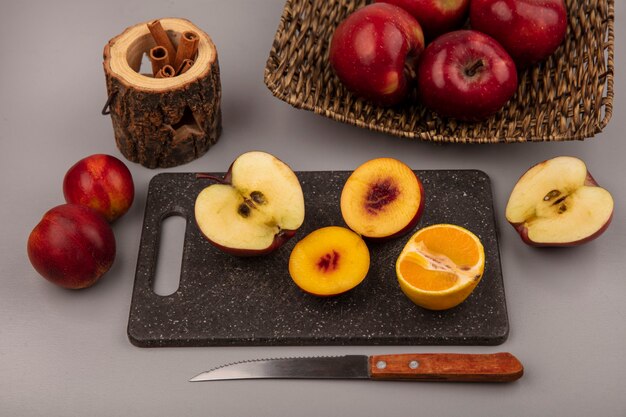 灰色の背景の枝編み細工品トレイにタンジェリンと赤いリンゴとナイフでリンゴと黒のキッチンボード上の新鮮な半分の桃の上面図