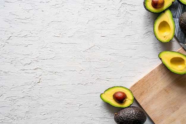 Top view of fresh halved avocados and a wooden cutting board on a white surface, copy space