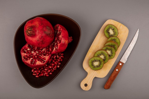 Top view of fresh halved and whole pomegranates on a bowl with slices of kiwi on a wooden kitchen board with knife