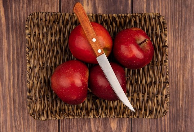 Вид сверху свежих сочных и красных яблок на плетеном подносе с ножом на деревянном фоне