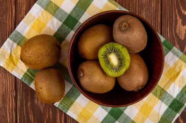 Top view of fresh kiwi fruits in a wooden bowl on plaid table napkin on wood