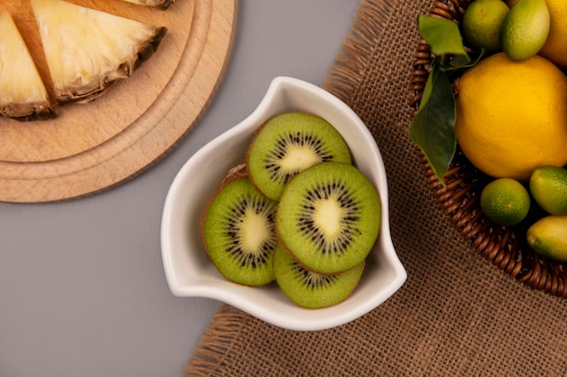 Top view of fresh kiwi slices on a bowl with fruits such as kinkans and lemons on a bucket on a sack cloth on a grey surface