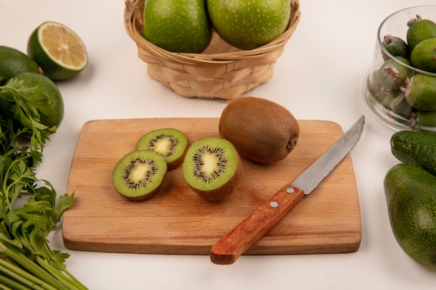 Top view of fresh kiwis on a kitchen board with knife with apples on a bucket with feijoas on a glass bowl with limes and avocados isolated on a white wall