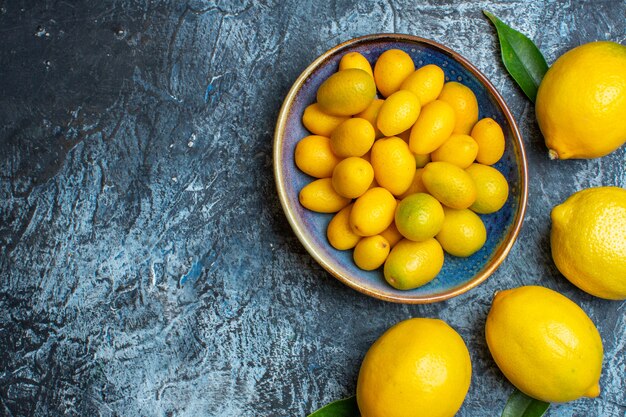Top view of fresh kumquats on a blue plate and natural lemons on the left side on dark background