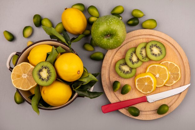 Top view of fresh lemons on a bowl with kiwi and lemon slices on a wooden kitchen board with knife with kinkans and apple isolated on a white wall