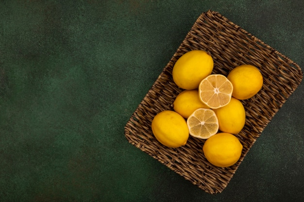 Вид сверху свежих лимонов на плетеном подносе на зеленом фоне с копией пространства