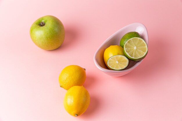 Вид сверху свежих лимонов с нарезанным лаймом внутри тарелки на светло-розовой поверхности