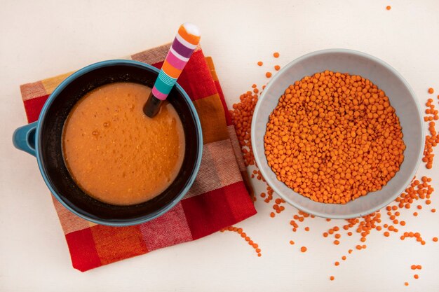 Top view of fresh lentils on a bowl with orange lentil soup on a bowl on a checked cloth with spoon on a white wall