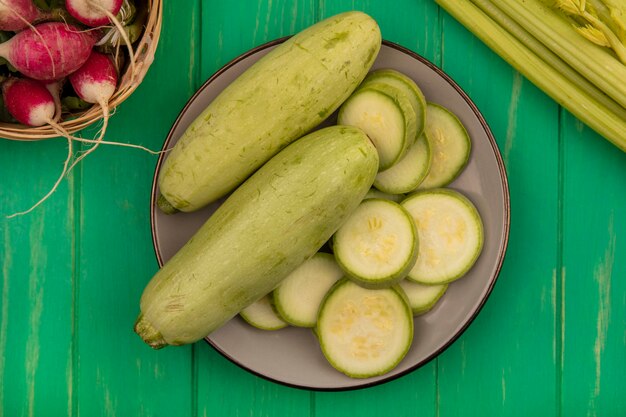 Top view of fresh light green zucchinis on a plate with radishes on a bucket with celery isolated on a green wooden wall