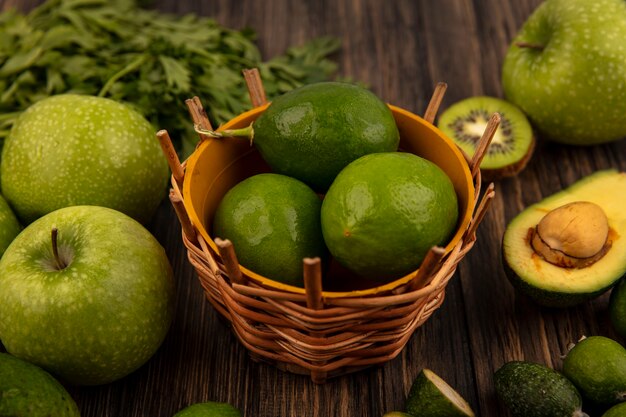 Top view of fresh limes on a bucket with green apples kiwi feijoas avocados and parsley isolated on a wooden wall