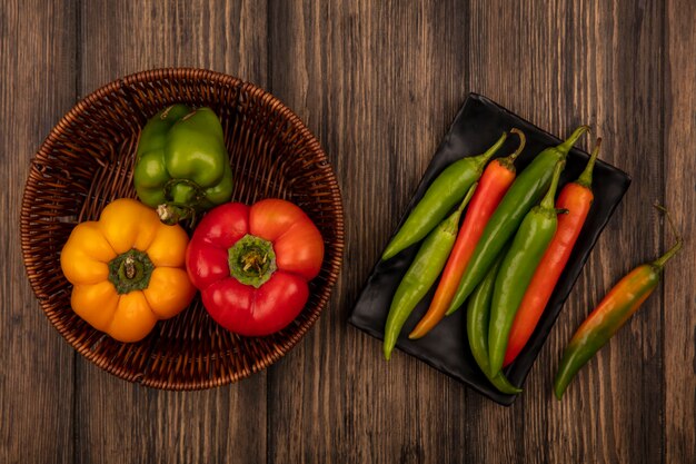 Top view of fresh long peppers on a black plate with bell peppers on a bucket on a wooden wall