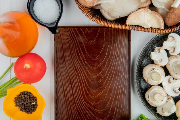 Top view of fresh mushrooms in a wicker basket and tomato bottle of olive oil salt and peppercorns arranged around wood board on white