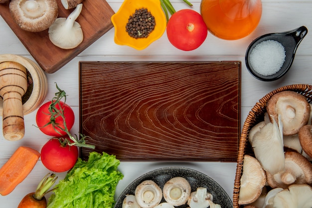 Top view of fresh mushrooms in a wicker basket and tomatoes bottle of olive oil salt and peppercorns arranged around wood board on white